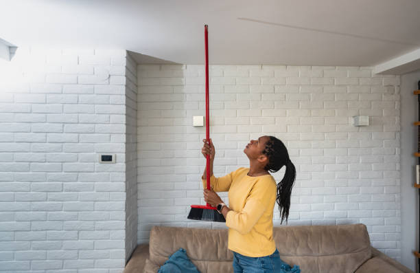 Женщина радостно чистит красной метлой паутину с белого кирпичного потолка светлой комнаты.