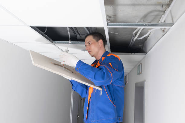 Техник в оранжево-синей форме осматривает или ремонтирует систему кондиционирования воздуха в потолочной панели.