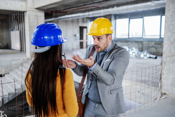 Два профессионала-строителя, мужчина и женщина в касках, беседуют внутри строящегося здания.